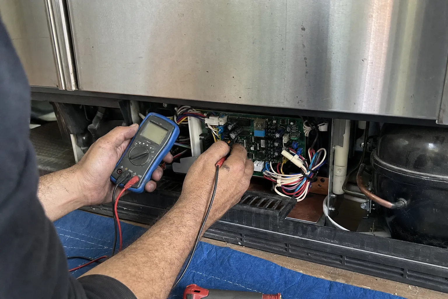 Multimeter probes on an exposed control board and wiring harness in the machine compartment under a stainless unit